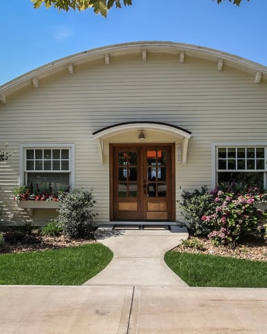 Cream-colored single-story house with arched entryway, double wooden doors, two windows, and pink flowering shrubs flanking the entrance