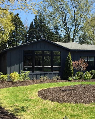 Modern black ranch house with large windows, surrounded by landscaped yard with mulch beds and young shrubs