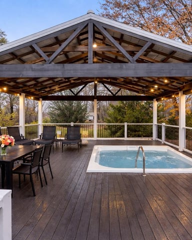 Large covered deck with hot tub, dining table, lounge chairs, and metal railings surrounded by autumn trees