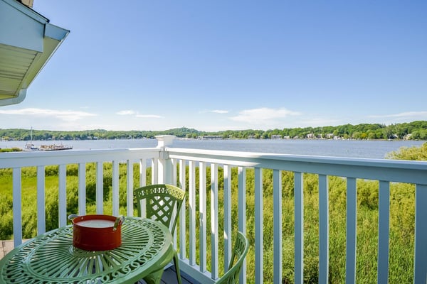 Waterfront deck with white railing, green metal table and chair, red pot, overlooking wide lake with boats and tree-lined shore