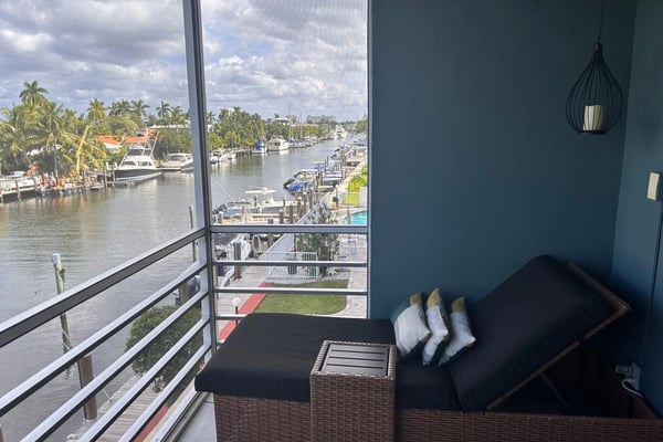 Waterfront balcony with wicker seating overlooking marina, moored boats, palm trees, and cloudy sky