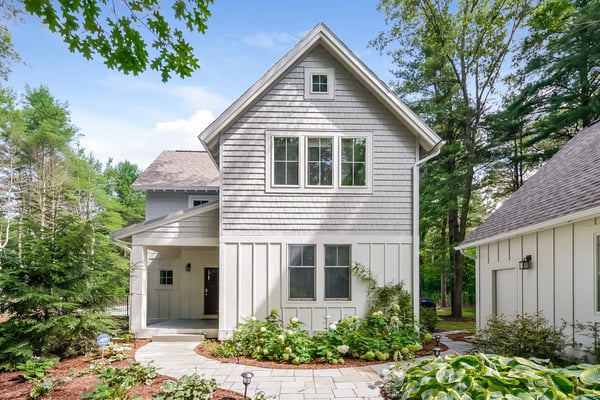 Modern farmhouse with gray shingle siding, white trim, and gabled roof surrounded by mature trees and landscaped front yard.