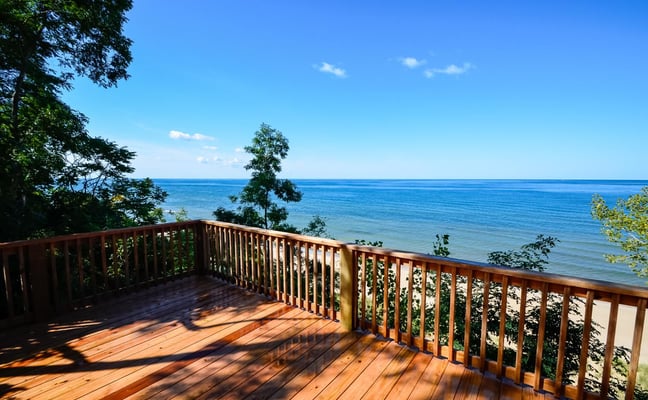 Wooden deck with railing overlooking calm blue lake and tree-lined shore under clear sky