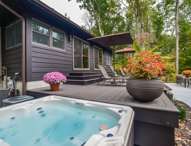 Modern dark cabin with hot tub, composite deck, potted flowers, umbrellas, and lounge chairs surrounded by tall trees